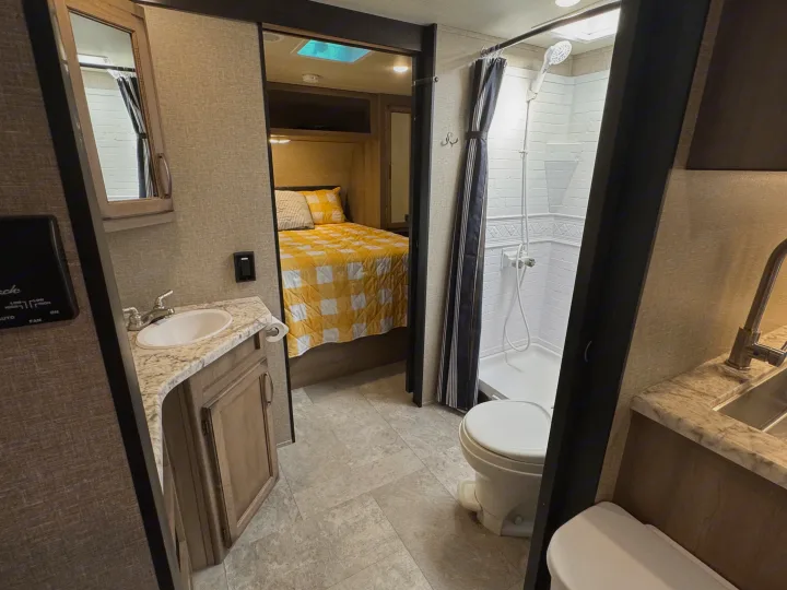 RV rental interior: A compact bathroom with sink, shower, and toilet, leading to a cozy bedroom with a yellow quilt.. Grand Design Transcend Xplor 2020