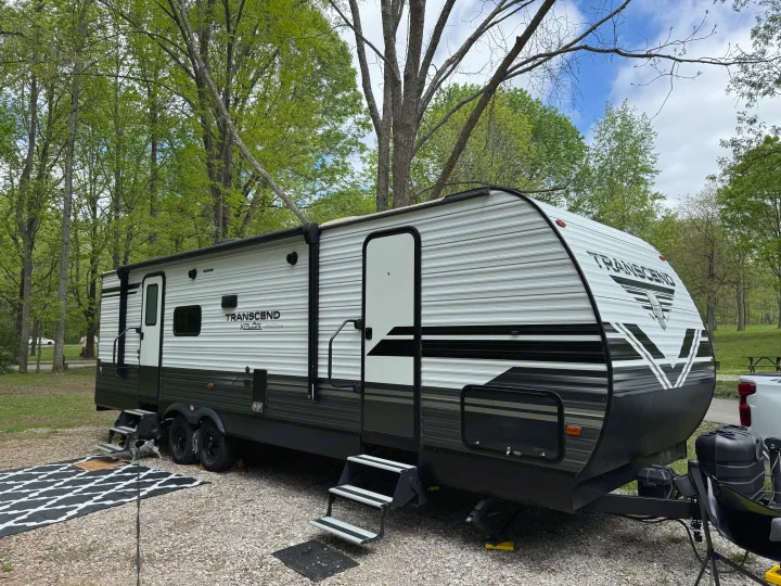 A modern Transcend Xplor travel trailer parked on a gravel site in a leafy campground, ready for an RV rental getaway.. Grand Design Transcend Xplor 2020