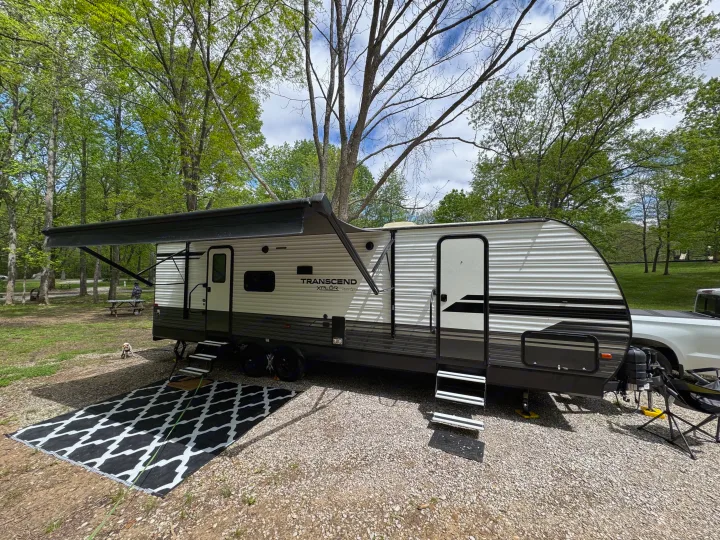 This RV rental travel trailer is set up at a shaded campsite with an awning, outdoor rug, and a cute dog. Perfect for camping!. Grand Design Transcend Xplor 2020