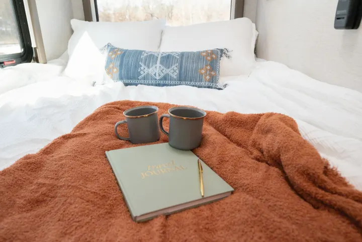 Cozy RV rental bed with fluffy blanket, travel journal, and mugs, perfect for planning adventures on the road.. Thor Motor Coach Eddie Bauer 21EBF 2025