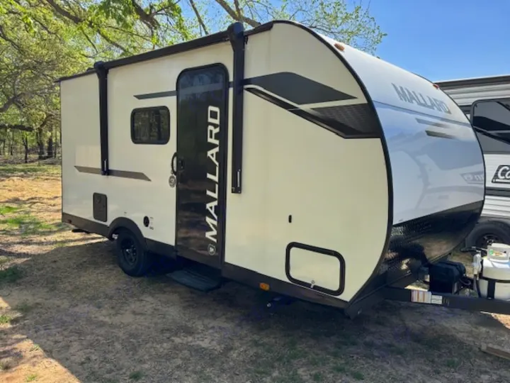 Cream-colored Mallard travel trailer for rv rental, parked outdoors. Features black accents & rolled awning.. Heartland Mallard Mallard 2025