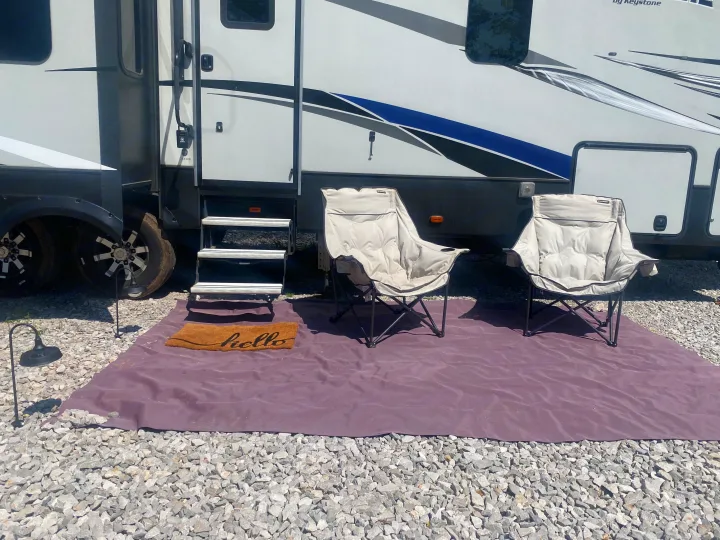 This RV rental features a comfortable outdoor setup with two padded chairs, a purple mat, and a 'hello' doormat on gravel.. Keystone RV Avalanche 2017