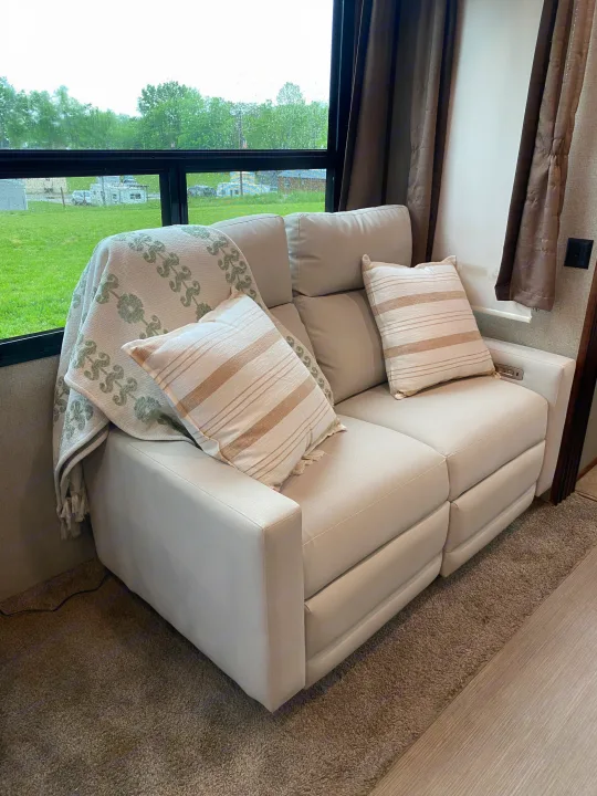Cozy RV rental interior with a light sofa, blanket, striped pillows, and a window view of a rainy green campground.. Keystone RV Avalanche 2017