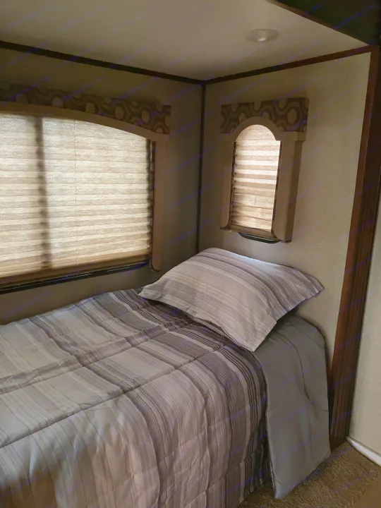 An RV rental bedroom features a striped bed, pillow, and two windows with drawn pleated blinds.. Keystone RV Avalanche 2017