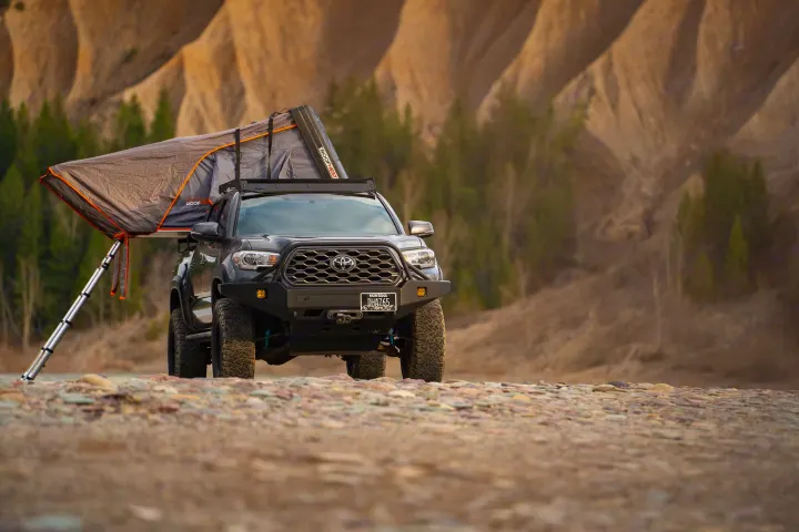 A dark gray Tacoma truck with an open rooftop tent sits on rocky ground, hills behind. Ideal for a rugged rv rental camping trip.. Toyota Tacoma TRD Off Road 2017