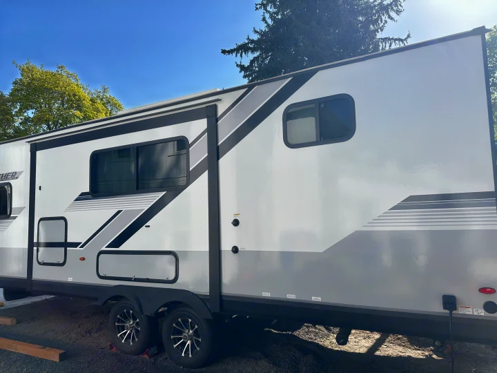 Modern white RV rental travel trailer with grey and black graphics, parked outdoors. Clear sky and trees visible.. Jayco Jay Feather 2024