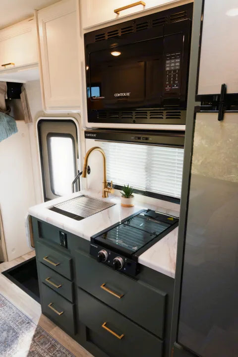 This RV rental kitchen has dark green cabinets, a marble-look counter, gold faucet, sink, and integrated microwave & stovetop.. Thor Motor Coach Eddie Bauer 21EBF 2025