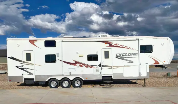 A white "Cyclone" fifth-wheel RV rental with brown decals, parked on gravel under a cloudy sky, seen in full length.. Heartland RVs Cyclone Toy Hauler 2008
