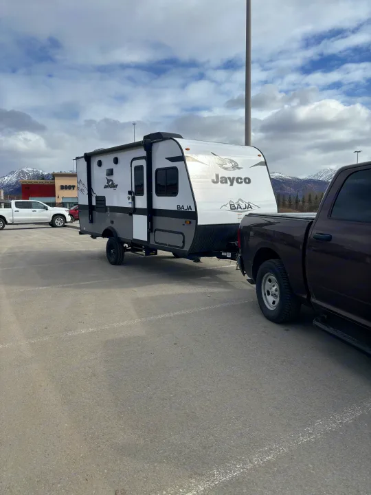 A dark pickup truck tows a white Jayco Baja RV rental travel trailer in a parking lot, with snowy mountains behind.. Jayco Jay Flight SLX Baja Edition 2022