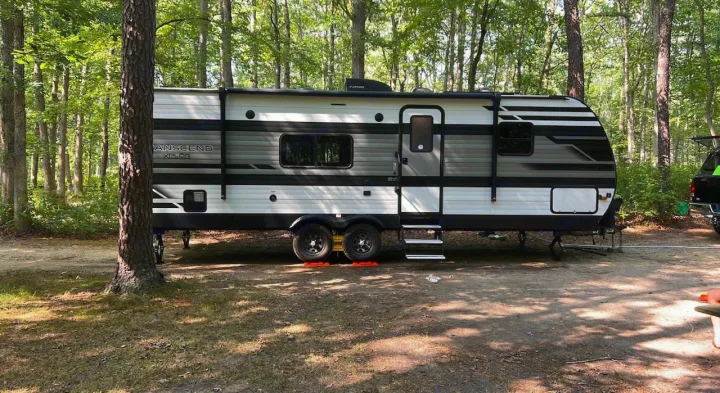An rv rental travel trailer, white and gray, is parked with leveling blocks at a shaded forest campsite, perfect for camping.. Grand Design Transcend Xplor 2021