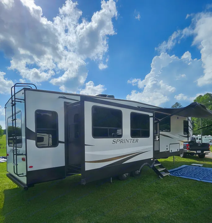 A large white & dark grey Sprinter RV rental is set up on green grass under a blue sky, featuring extended slide-outs.. Keystone Sprinter 2022