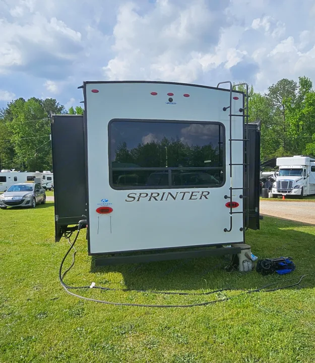 Rear view of a white Sprinter RV, possibly an rv rental, at a grassy campsite with power hookups and a ladder.. Keystone Sprinter 2022