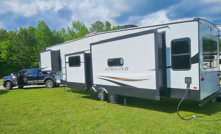 Modern fifth-wheel RV rental with slide-outs extended, parked on grass next to its tow truck under a cloudy sky.. Keystone Sprinter 2022
