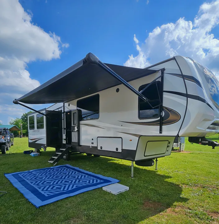 Large fifth-wheel RV rental with awning extended and mat on green grass. Perfect for outdoor camping under a sunny sky.. Keystone Sprinter 2022