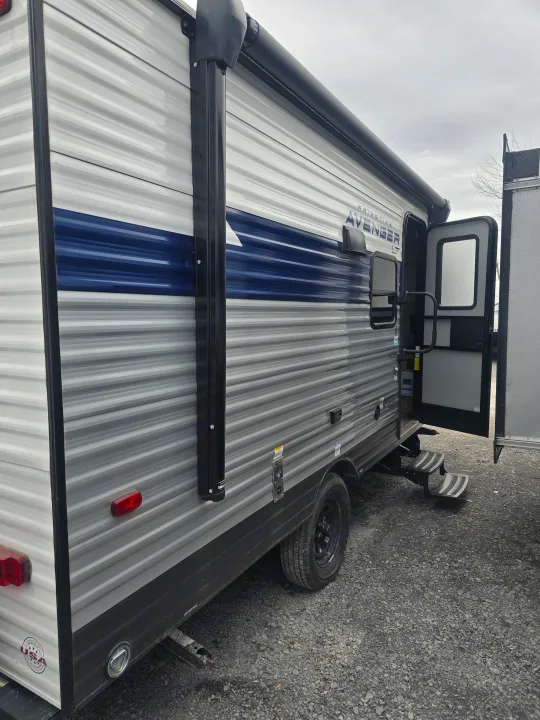 This modern grey & blue Primetime Avenger LT travel trailer, with open door & steps, is ready for RV rental.. Prime Time Avenger LT 16BH 2023