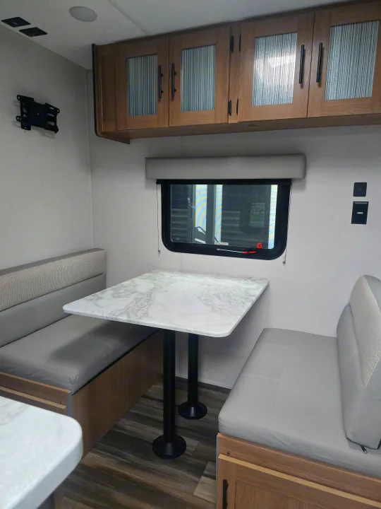 This RV rental interior features a modern dinette with a marble-look table, grey benches, and wood overhead cabinets.. Prime Time Avenger LT 16BH 2023