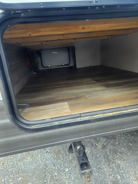 Spacious under-bed storage inside an RV rental, featuring wood-look floor, access hatch, and a visible stabilizer jack.. Prime Time Avenger LT 16BH 2023