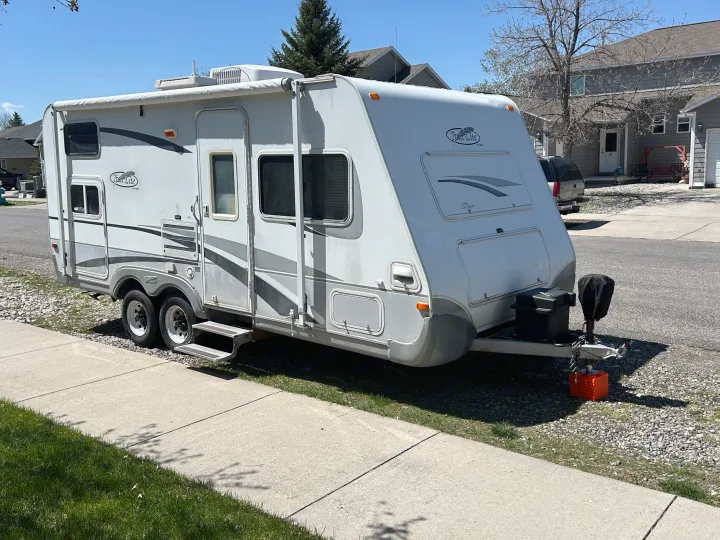 A white Trail Lite travel trailer RV rental, with dual axles, is parked on gravel by a sidewalk on a sunny day.. R-Vision Trail-Lite 2005