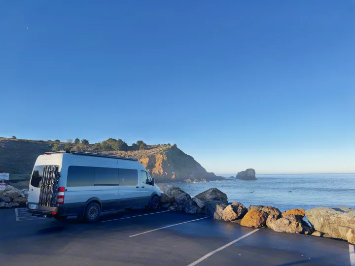 An RV rental van enjoys a scenic coastal park overlooking the ocean, with surfers, cliffs, and a crescent moon.. Mercedes-Benz Sprinter RV Motorhome Campervan 2012