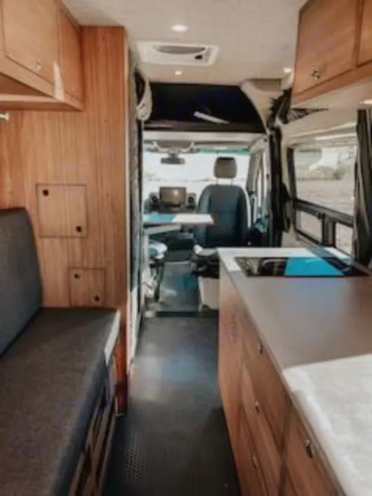 Interior of a modern RV rental camper van, showing kitchen, wood cabinets, bench seating, and front cab area.. Mercedes-Benz Sprinter RV Motorhome Campervan 2020