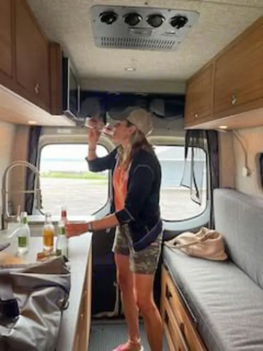 A woman enjoys a drink in the kitchen area of a modern **RV rental**. Inside, a sink, microwave, and bench seating provide comfort for travel.. Mercedes-Benz Sprinter RV Motorhome Campervan 2020