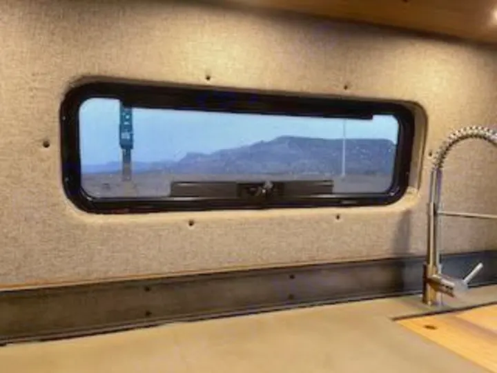 Inside an RV rental, a window offers a desert mountain view, with a kitchen sink visible nearby. Ideal for scenic road trips.. Mercedes-Benz Sprinter RV Motorhome Campervan 2020
