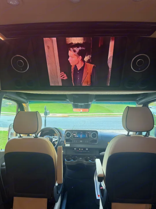 Spacious RV rental interior with a large screen playing a movie above the dashboard. Comfortable travel and entertainment setup.. Mercedes-Benz Sprinter 2025