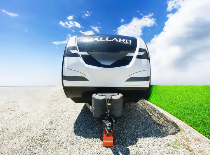 This RV rental, a white and black Mallard trailer, is ready for travel. Parked on gravel, with unusual green grass.. Heartland RVs Mallard 2021