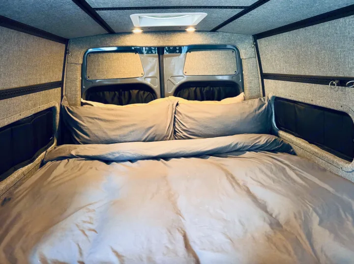 Inside an RV rental camper van: A neatly made bed with grey bedding and textured wall/ceiling panels.. Mercedes-Benz Sprinter 2022