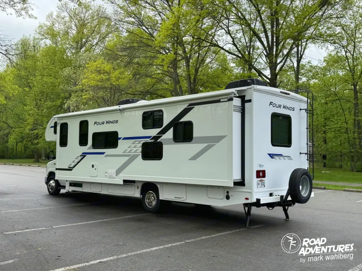 A white "Four Winds" RV rental with an extended slide-out, parked in a lot surrounded by green trees, ready for travel.. Thor Motor Coach Four Winds 2026