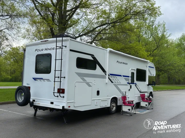 This Four Winds RV rental is parked among lush trees, with camping chairs set up for a relaxing outdoor stop.. Thor Motor Coach Four Winds 2026