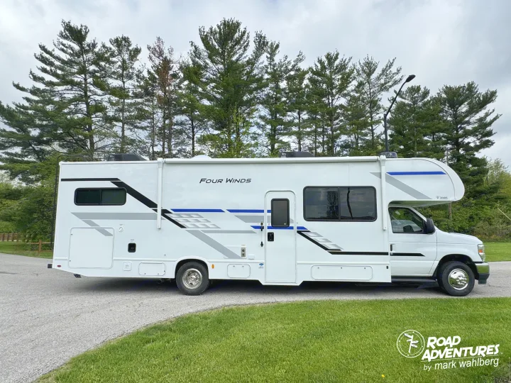 This white Four Winds Class C RV rental, with blue & black graphics, is parked on an asphalt road surrounded by tall pine trees.. Thor Motor Coach Four Winds 2026