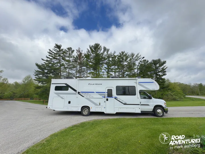 This Four Winds RV rental is ready for a road adventure, parked on a drive near tall pine trees under a cloudy sky.. Thor Motor Coach Four Winds 2026
