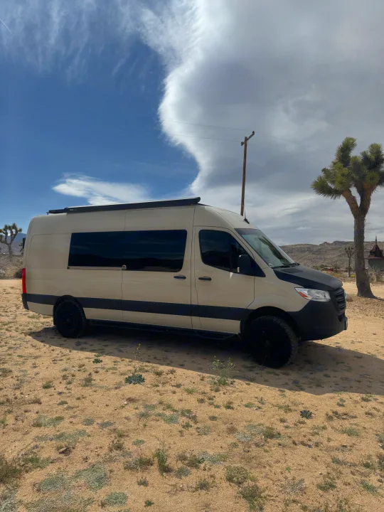 Tan Mercedes Sprinter RV rental van with off-road tires, parked in a desert landscape featuring Joshua trees and clouds.. VanCraft VanCraft Class B 2022