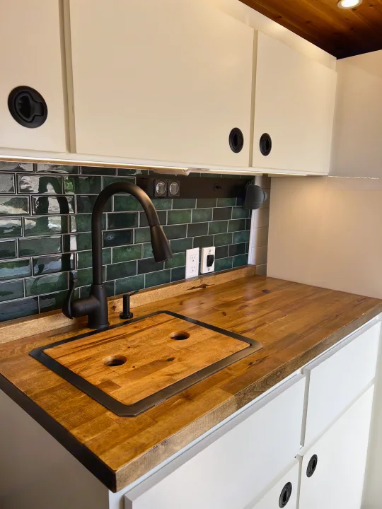 Modern RV rental kitchenette in a camper. Features a dark green tile backsplash, wood counter, black faucet, white cabinets.. Dodge Ram Promaster 2500 2022