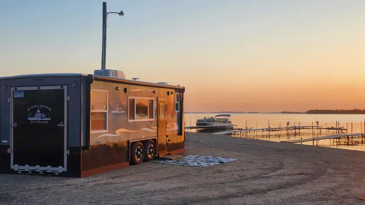 A gray Ice Castle Toy Hauler RV is parked on a sandy shore at sunset, with a pontoon boat and docks on the lake. Perfect rv rental.. Ice Castle Fish Houses Toy Hauler 2022