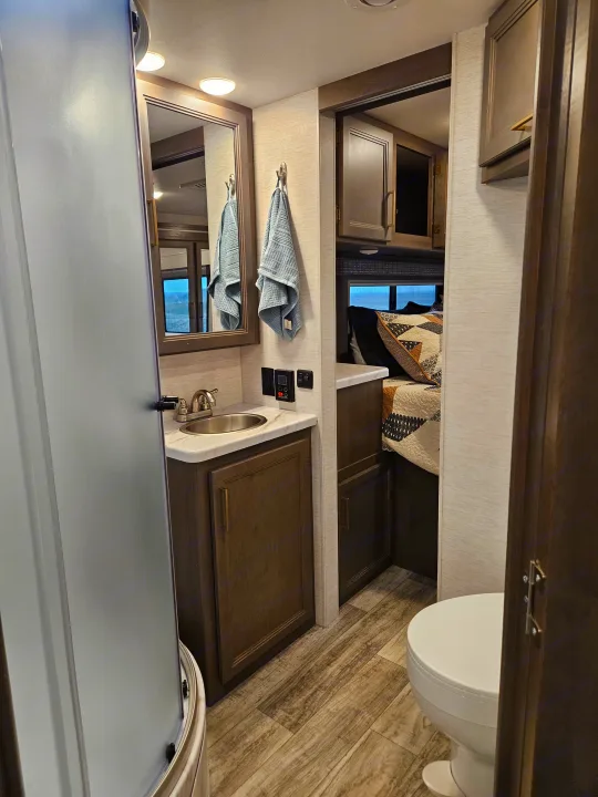 RV rental interior: A compact, modern bathroom with sink, shower, and toilet, leading to a cozy bedroom area.. Thor Chateau 2024