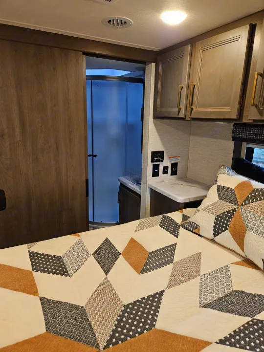 RV rental: Bedroom with geometric quilt, wood cabinets, and an open sliding door to a modern bathroom with a glass shower.. Thor Chateau 2024