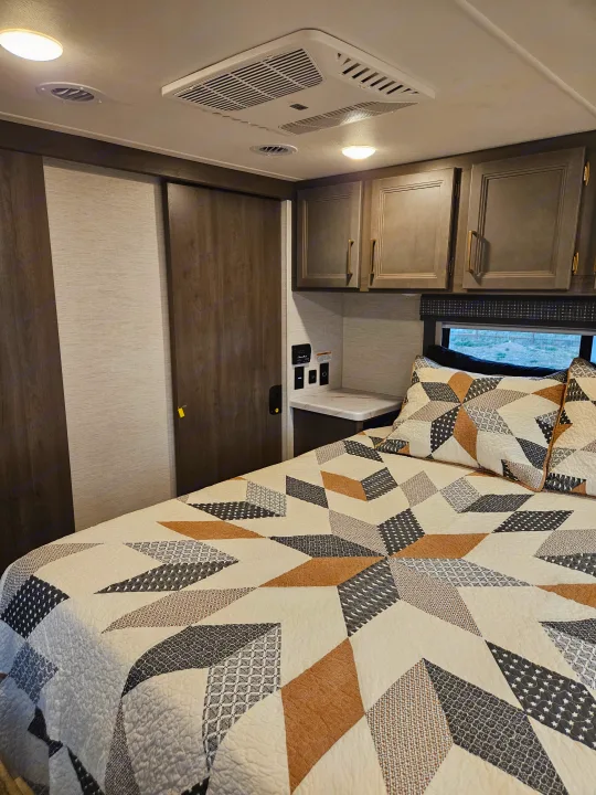 Modern RV rental bedroom featuring a bed with a geometric quilt, overhead cabinets, and built-in controls.. Thor Chateau 2024