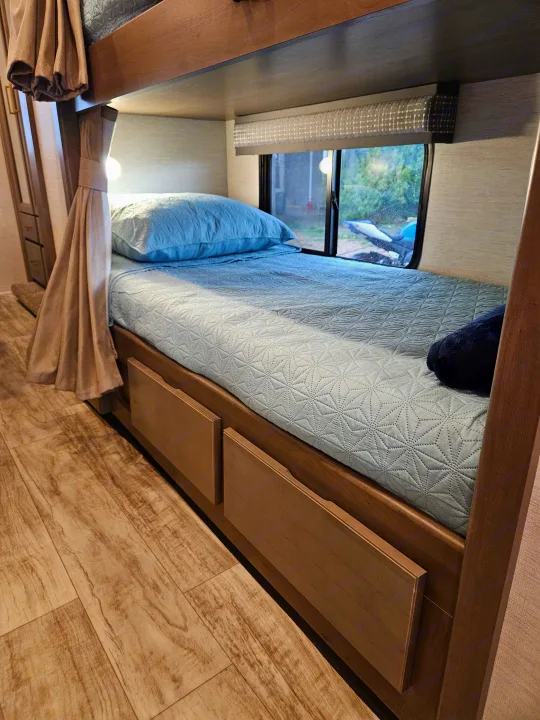 Comfortable RV rental lower bunk bed: blue quilted bedding, window view, and built-in storage drawers.. Thor Chateau 2024