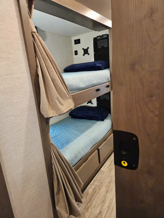 This RV rental features a cozy bunk bed area with light blue bedding, privacy curtains, outlets, and storage solutions.. Thor Chateau 2024