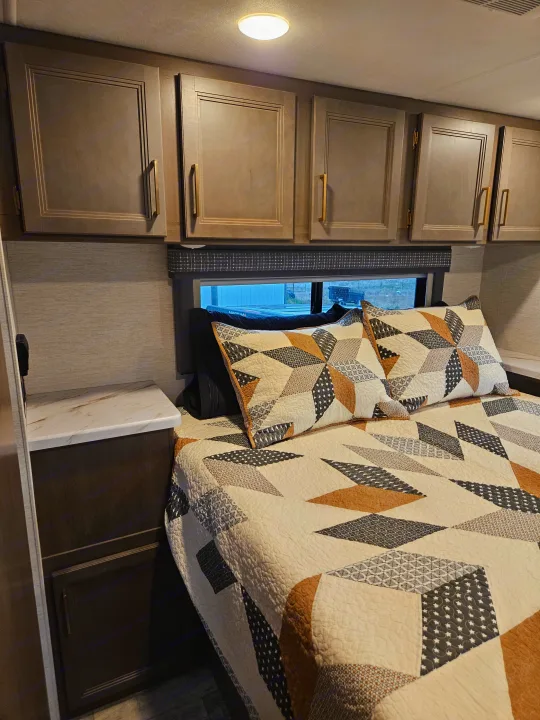 This RV rental bedroom features a geometric quilt, ample overhead cabinets, and a convenient nightstand, perfect for your travels.. Thor Chateau 2024