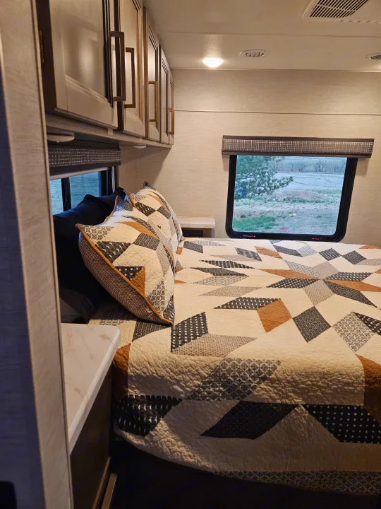 Inside an **RV rental**: a bright bedroom with a patterned quilt, convenient overhead storage, and a picturesque window view.. Thor Chateau 2024