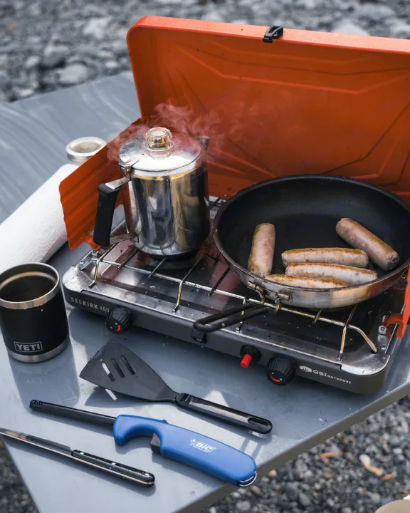 RV rental breakfast: Steaming coffee & sausages cook on a camp stove. A Yeti mug, spatula, and lighter are ready for outdoor adventures.. Ford Ford F150 2024