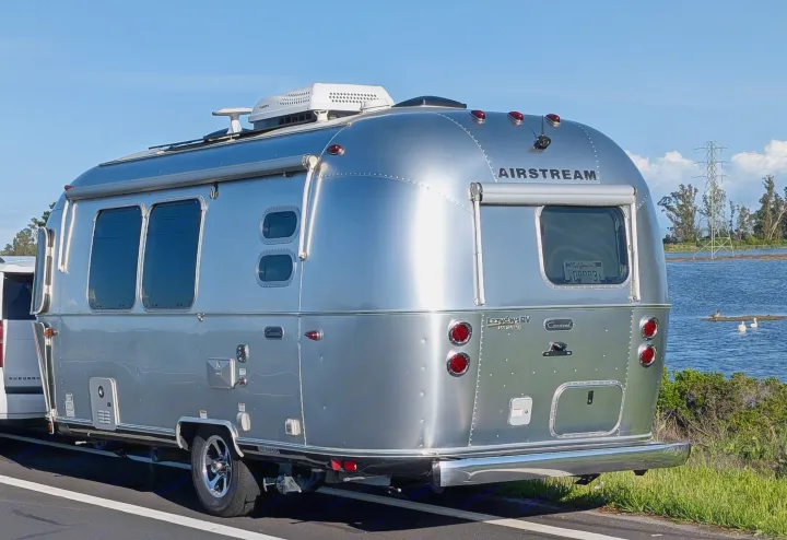 This sleek silver Airstream travel trailer, an excellent rv rental option, is parked by a scenic lake under blue skies.. Airstream Caravel 2023
