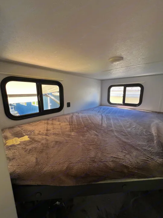 Bright RV rental interior showing a large, cozy bunk bed with a soft grey blanket, two windows, and textured ceiling.. Forest River Salem FSX 2021