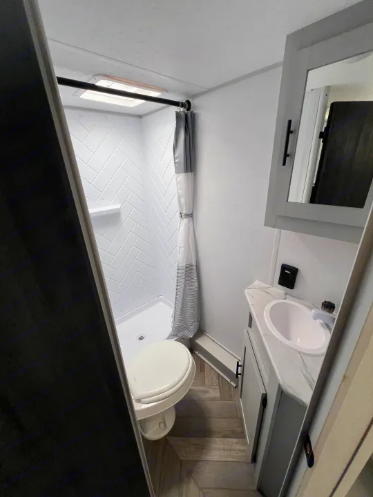 Compact RV rental bathroom featuring a shower with herringbone walls, toilet, and vanity with a sink. Modern gray and white interior.. Forest River Salem FSX 2021