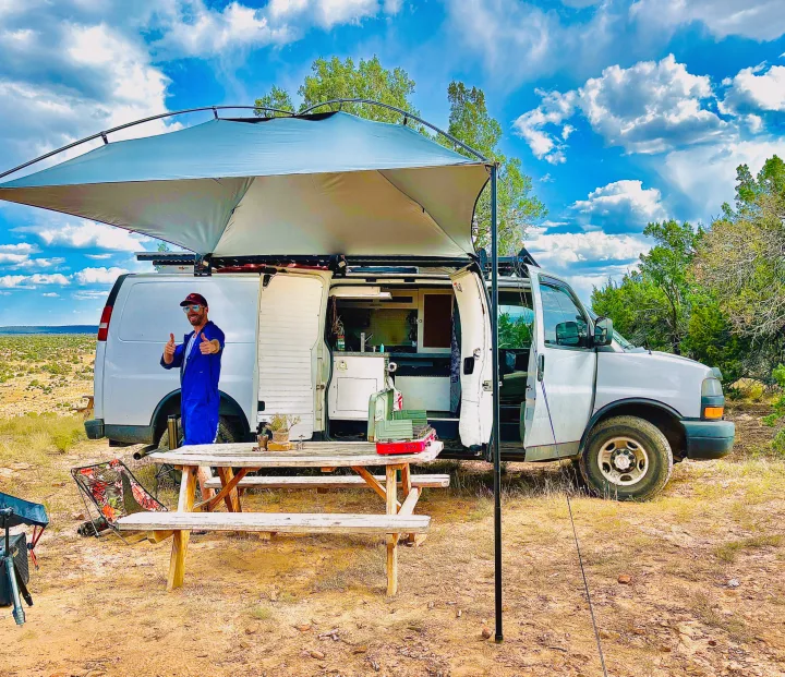 A man gives thumbs up by his van life setup with an awning and picnic table in a desert landscape, ideal for an rv rental adventure.. Chevrolet Express 3500 2012
