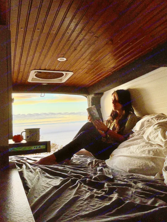 Cozy rv rental escape: Woman reads in bed, admiring a vibrant sunset and ocean view from her camper van.. Chevrolet Express 3500 2012