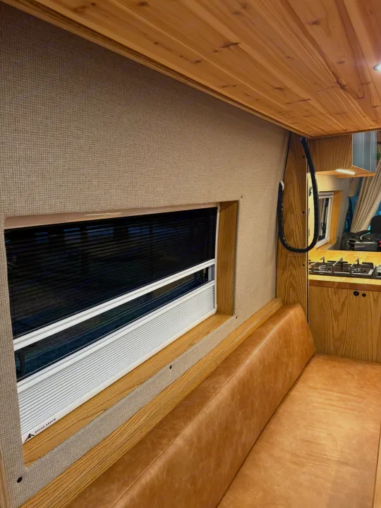 Inside an rv rental: inviting living space with wood finishes, comfy bench, window blinds, and kitchen area.. Ford Transit 2023
